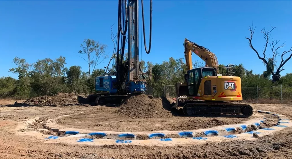 A drilling rig and a yellow CAT excavator work on a dirt construction site, with blue markings forming a circle on the ground. Trees and a fence are visible in the background.