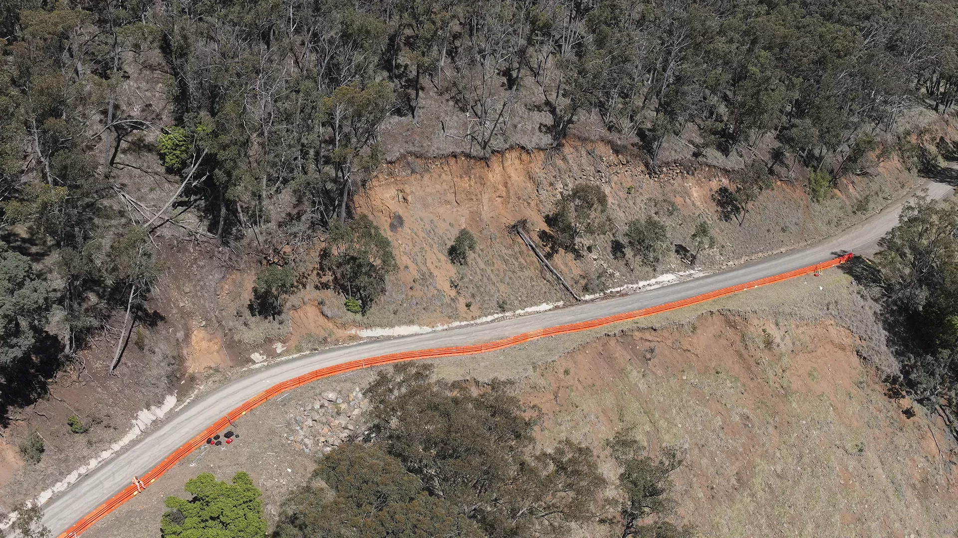 Aerial view of a narrow road with orange safety barriers along a steep, eroded hillside surrounded by sparse trees and vegetation.