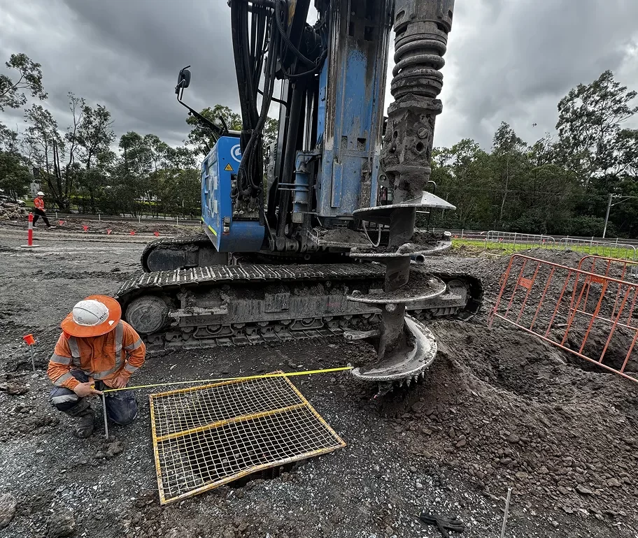 A construction worker measures near a large drilling machine at a muddy construction site, with orange safety barriers and cloudy skies in the background.