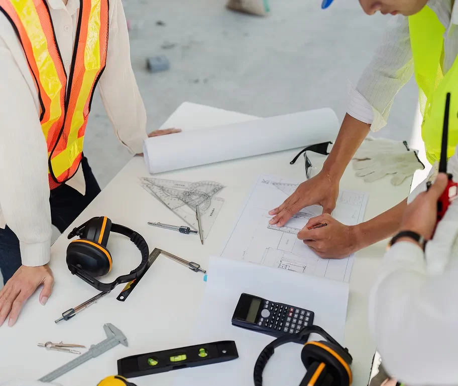 Three people in safety vests review building plans on a table covered with tools, blueprints, a calculator, ear protection, and a walkie-talkie.