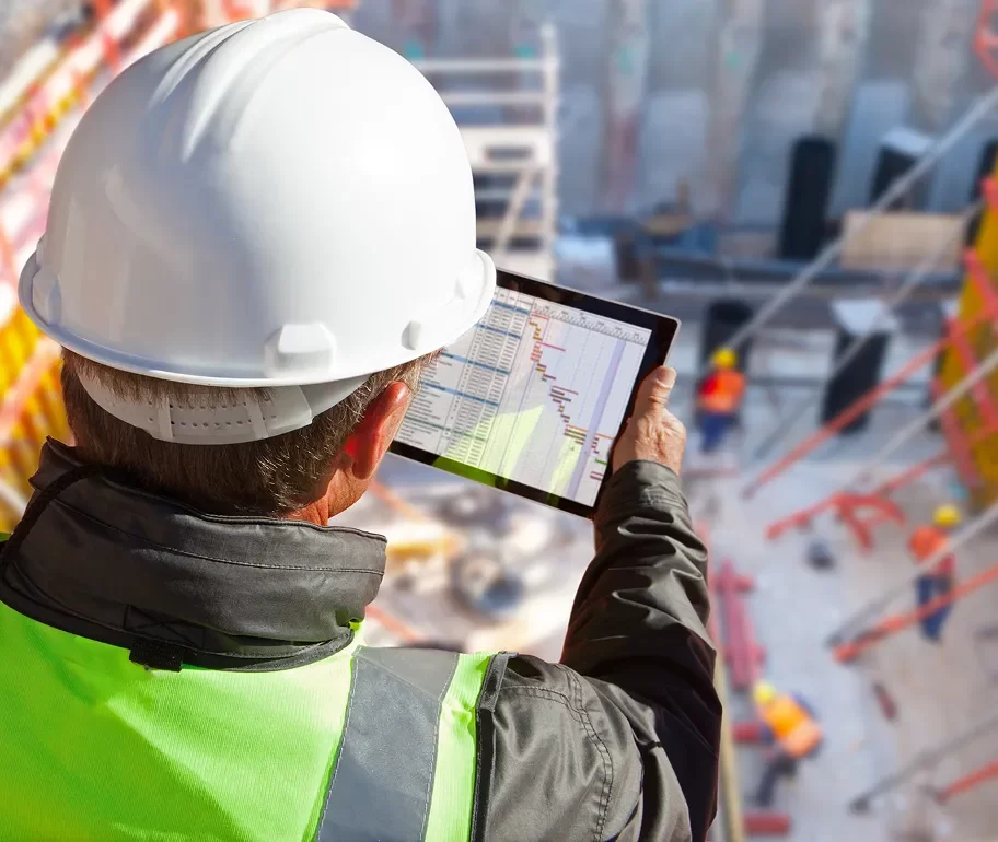 A construction worker in a hard hat and safety vest reviews a project schedule on a tablet at a building site.