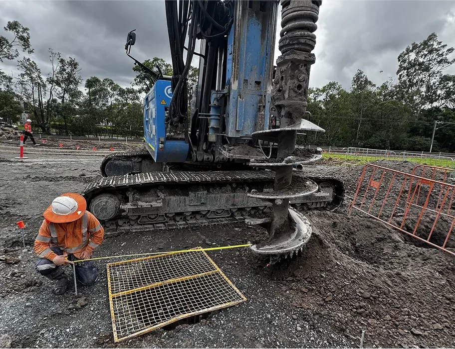 A construction worker measures the distance from a metal grate while a large drilling machine operates on a gravel surface near safety barriers and trees under a cloudy sky.