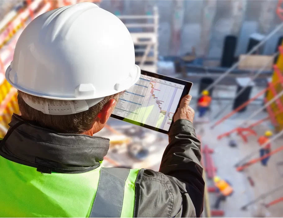A construction worker in a hard hat and safety vest checks a project schedule on a tablet at a building site.