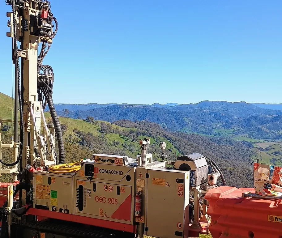 A drilling rig labeled "COMACCHIO GEO 205" is positioned on a hillside, with green valleys and distant mountains under a clear blue sky in the background.