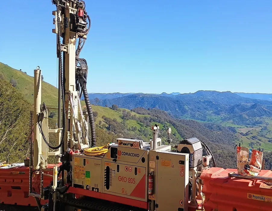 Drilling rig and equipment labeled "GEO 905" set up on a hillside, with mountains, green hills, and a clear blue sky in the background.