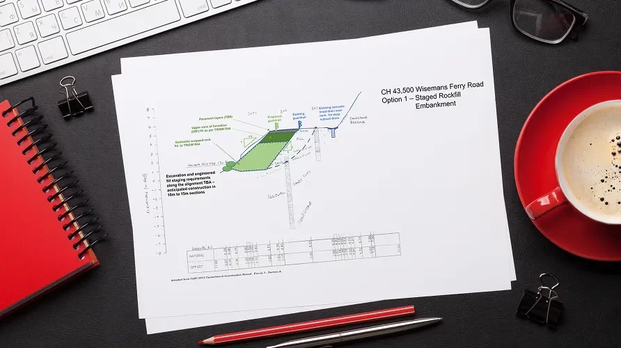A printed engineering diagram titled "CH 43.500 Wisemans Ferry Road Option 1 – Staged Rockfill Embankment" lies on a desk with a keyboard, notepad, pen, and a cup of coffee.