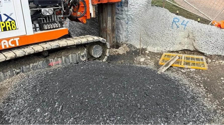 A construction machine with metal tracks is positioned next to a concrete retaining wall, with a pile of gravel and a yellow metal grate on the ground nearby.
