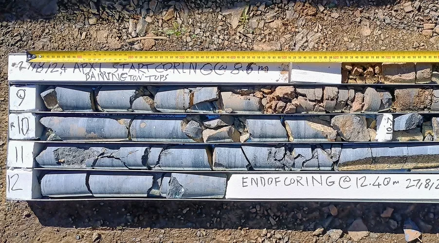 Three rows of cylindrical rock core samples in a labeled box, with a yellow measuring tape laid across the top for scale.
