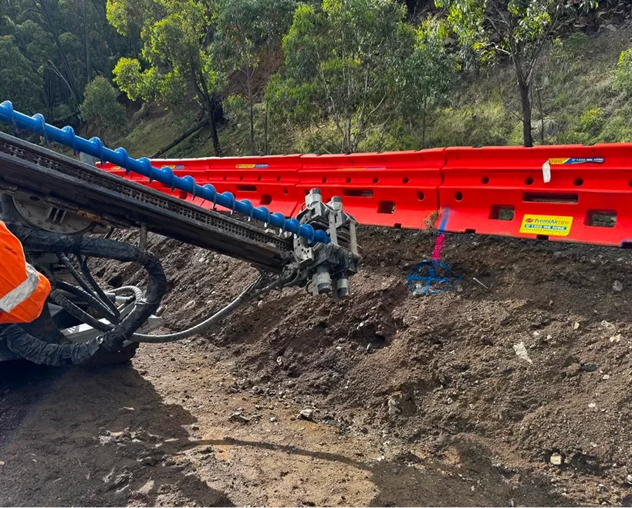 A construction machine drills into a dirt embankment beside a bright red highway guardrail, with trees and vegetation in the background.