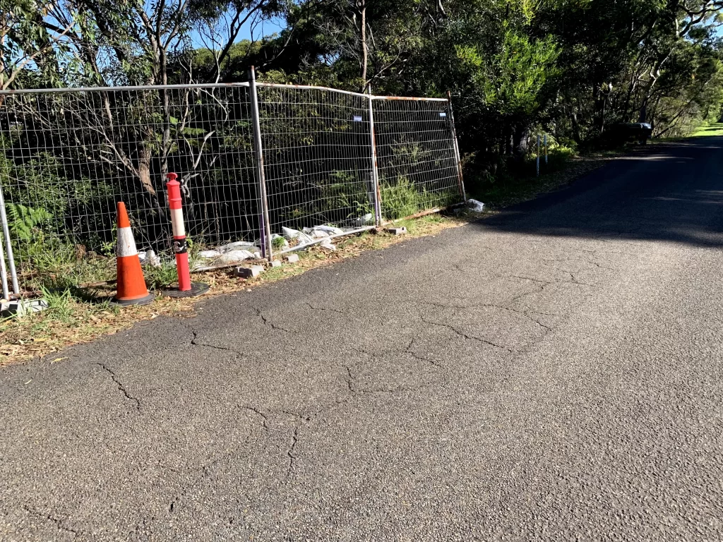 A wire fence with traffic cones and bollards runs alongside a cracked paved road, with trees and vegetation in the background.