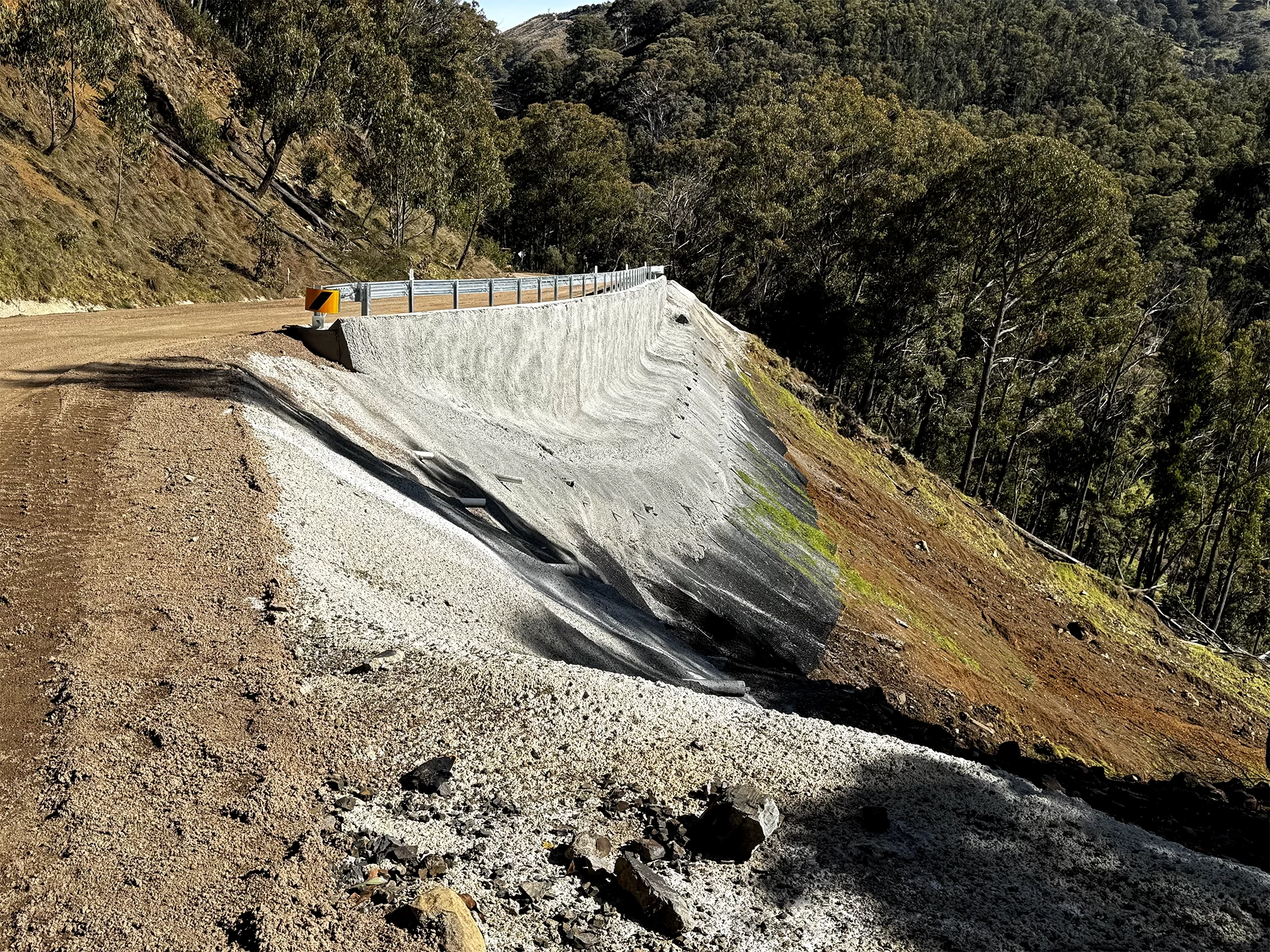 A gravel road runs alongside a concrete retaining wall on a slope, bordered by trees and dense vegetation on a hillside.