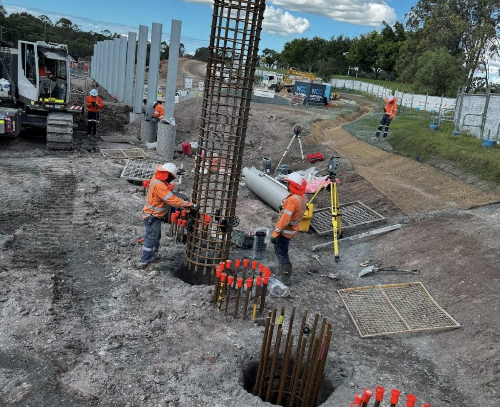 Construction workers install steel reinforcement cages into drilled holes at a roadside construction site, preparing for concrete pouring, with equipment and machinery present nearby.