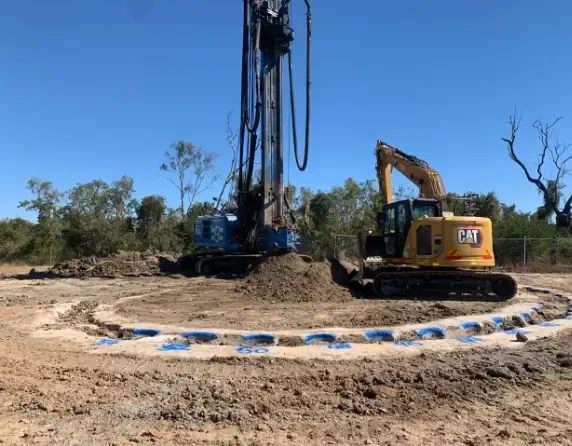 A yellow excavator and a blue piling rig operate on a dirt construction site, surrounded by marked circular lines, with trees and clear blue sky in the background.